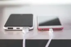 Zwei Smartphones, eines schwarz, eines rot, sind auf einer hellen Oberfläche mit weißen Ladekabeln verbunden.