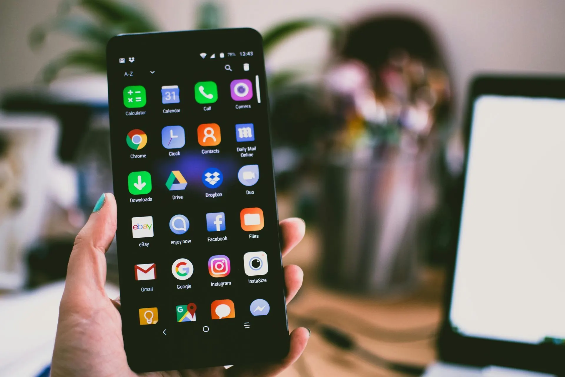 Eine Hand hält ein schwarzes Smartphone, auf dem ein Raster von App-Symbolen auf einem Schreibtisch angezeigt wird.