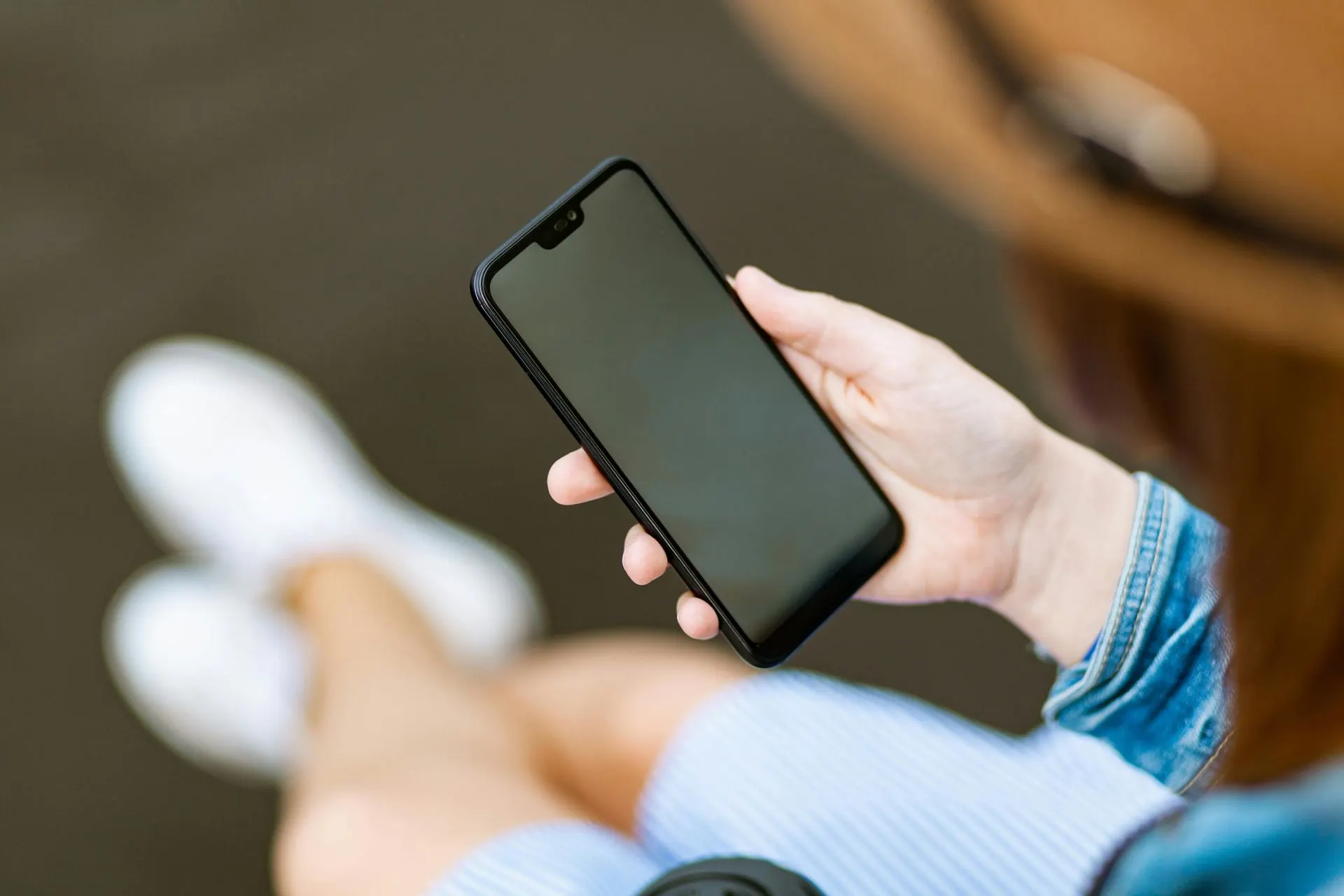 Eine sitzende Person hält ein schwarzes Smartphone mit leerem Bildschirm, trägt eine Jeansjacke.