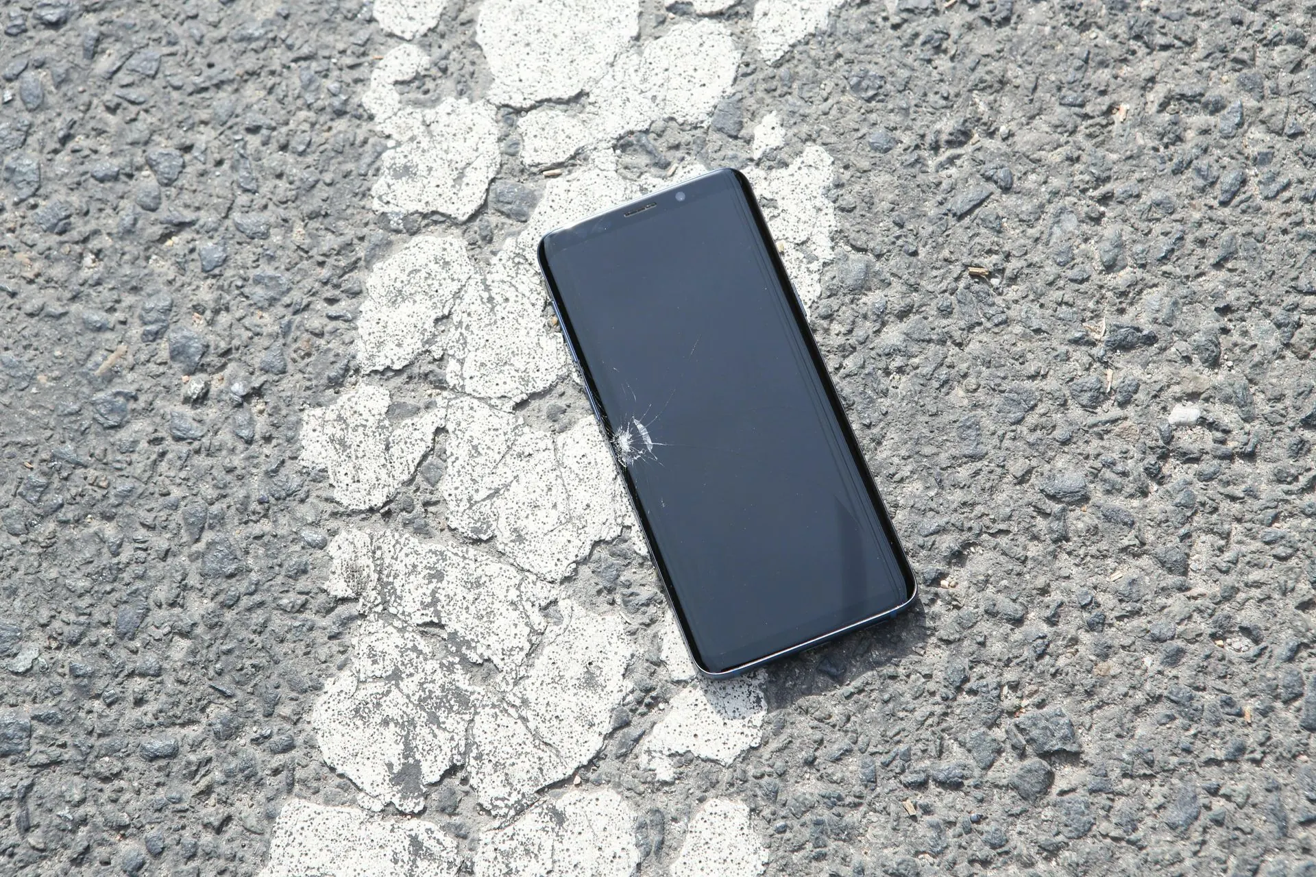 Schwarzes Smartphone liegt auf rissigem Asphalt mit einer weißen Linie.