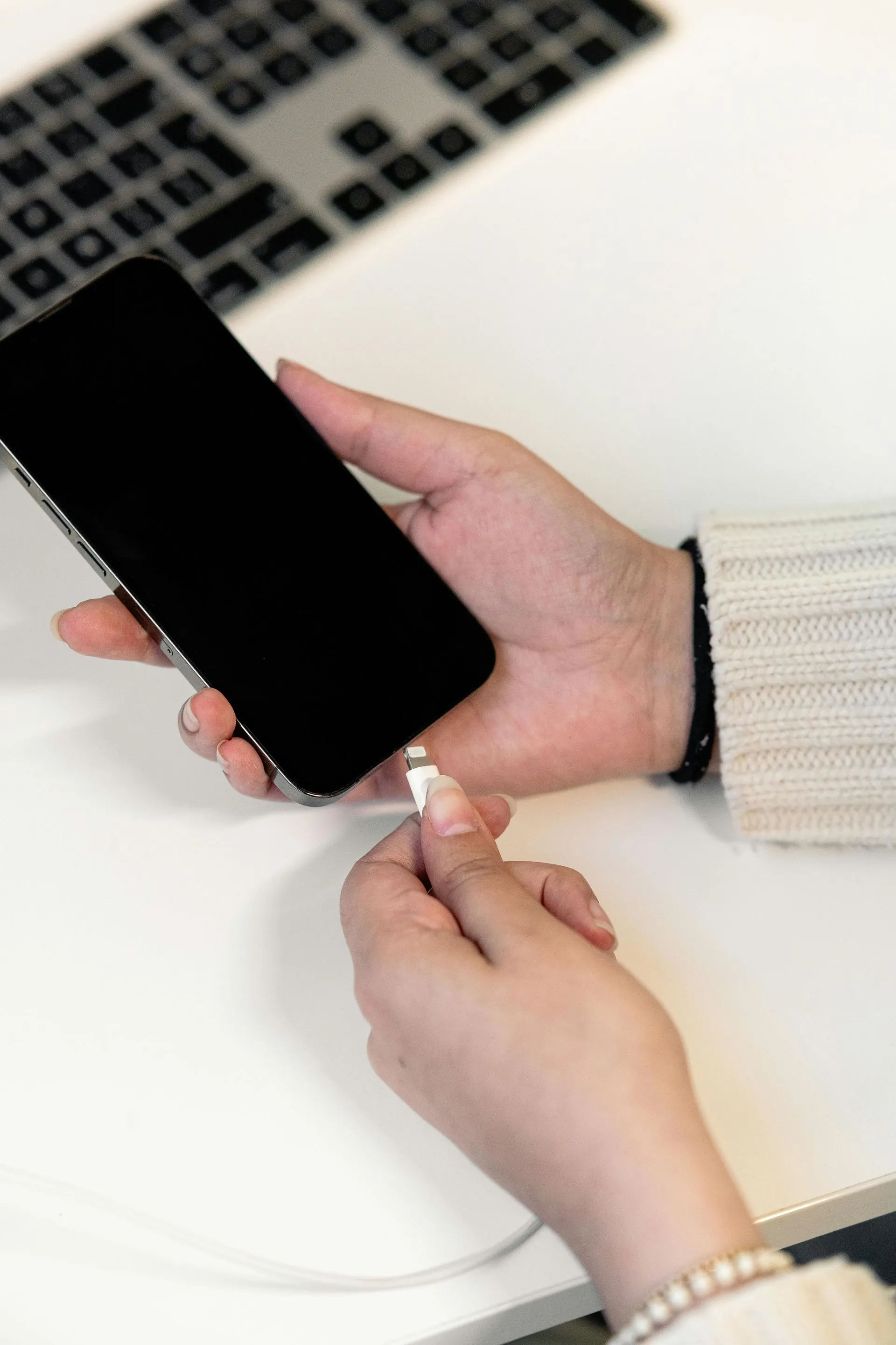 Eine Person steckt ein Ladekabel in ein schwarzes Smartphone auf einem weißen Schreibtisch; im Hintergrund ist eine Tastatur zu sehen.