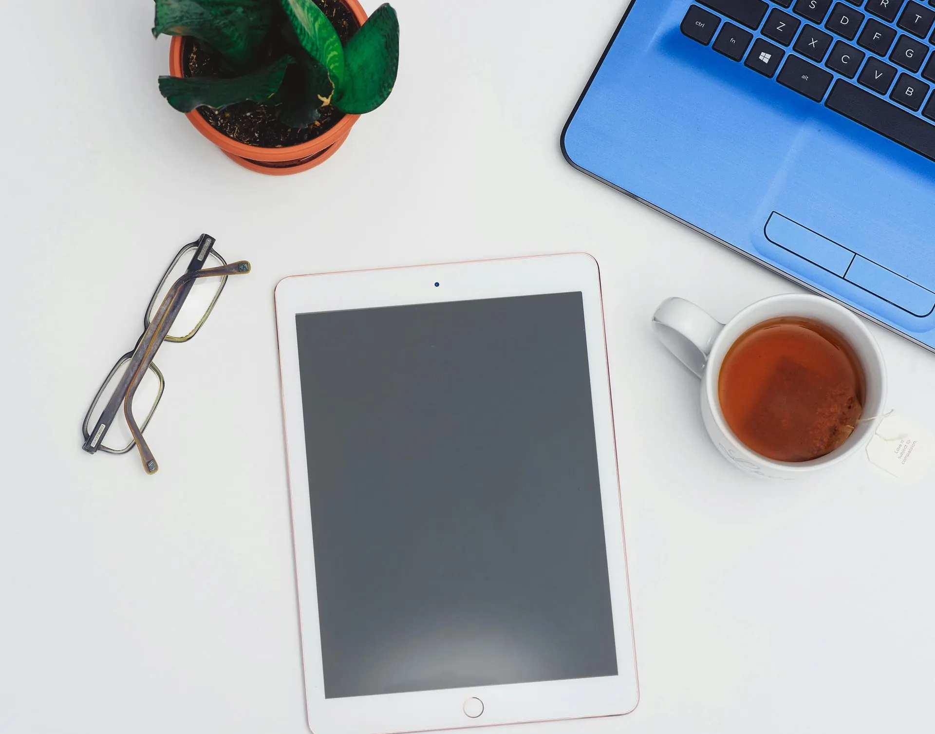 Weißer Schreibtisch mit Tablet, blauem Laptop, Brille, einer Pflanze und einer Tasse Tee.