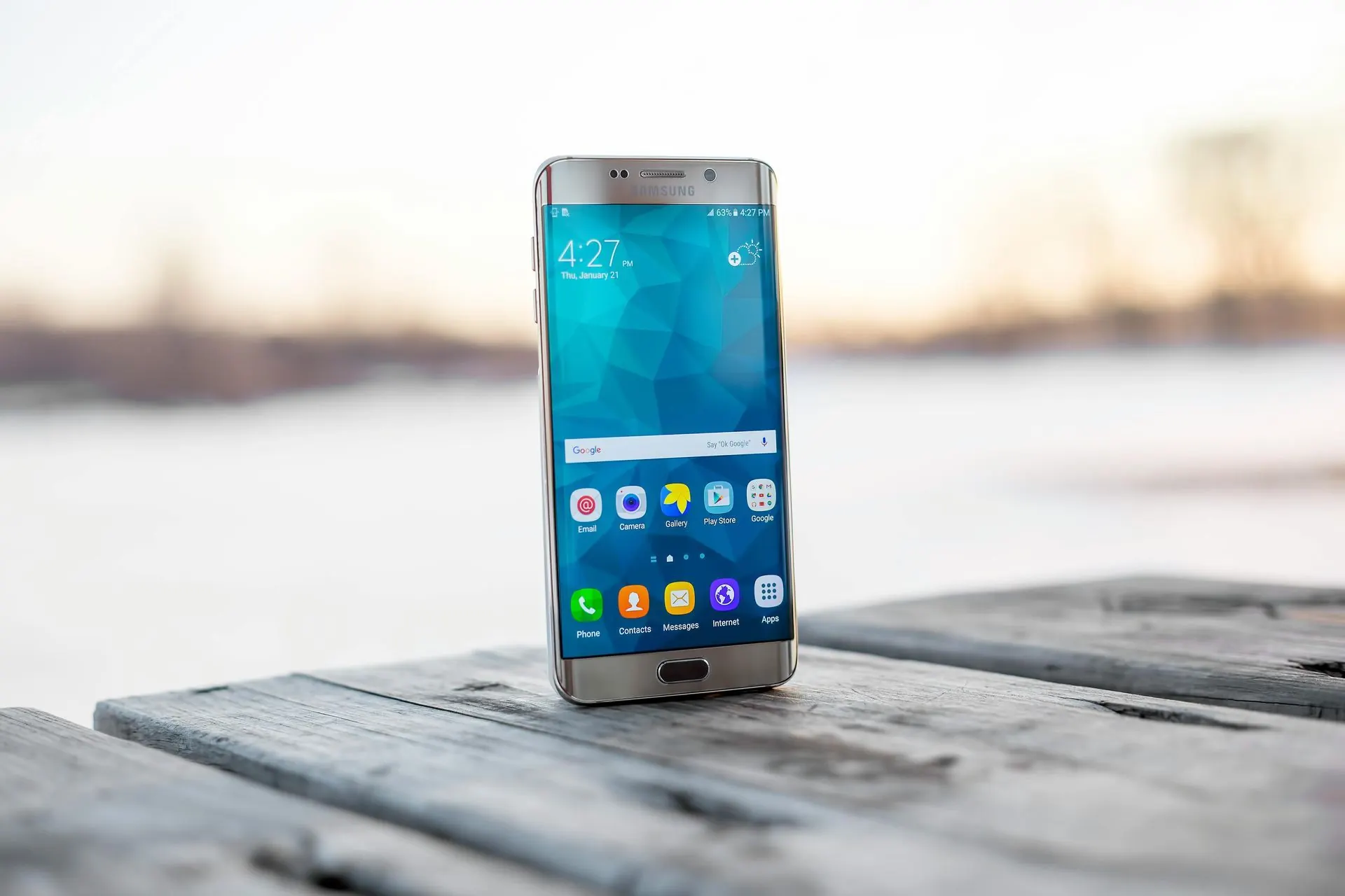 Silbernes Smartphone auf einer verwitterten Holzoberfläche, im Hintergrund eine verschwommene winterliche Außenkulisse.