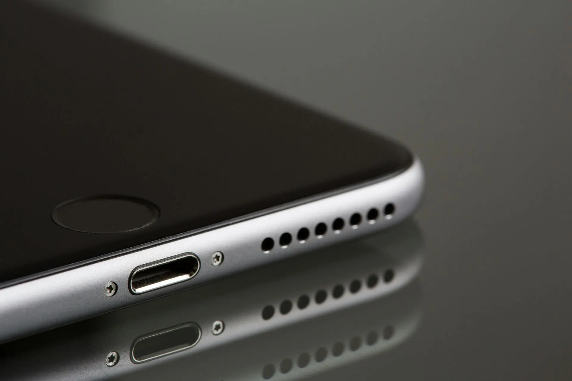 Nahaufnahme eines silbernen Smartphones, zu sehen sind der Ladeanschluss, die Lautsprecheröffnungen und dessen Spiegelung auf einer glänzenden Oberfläche.