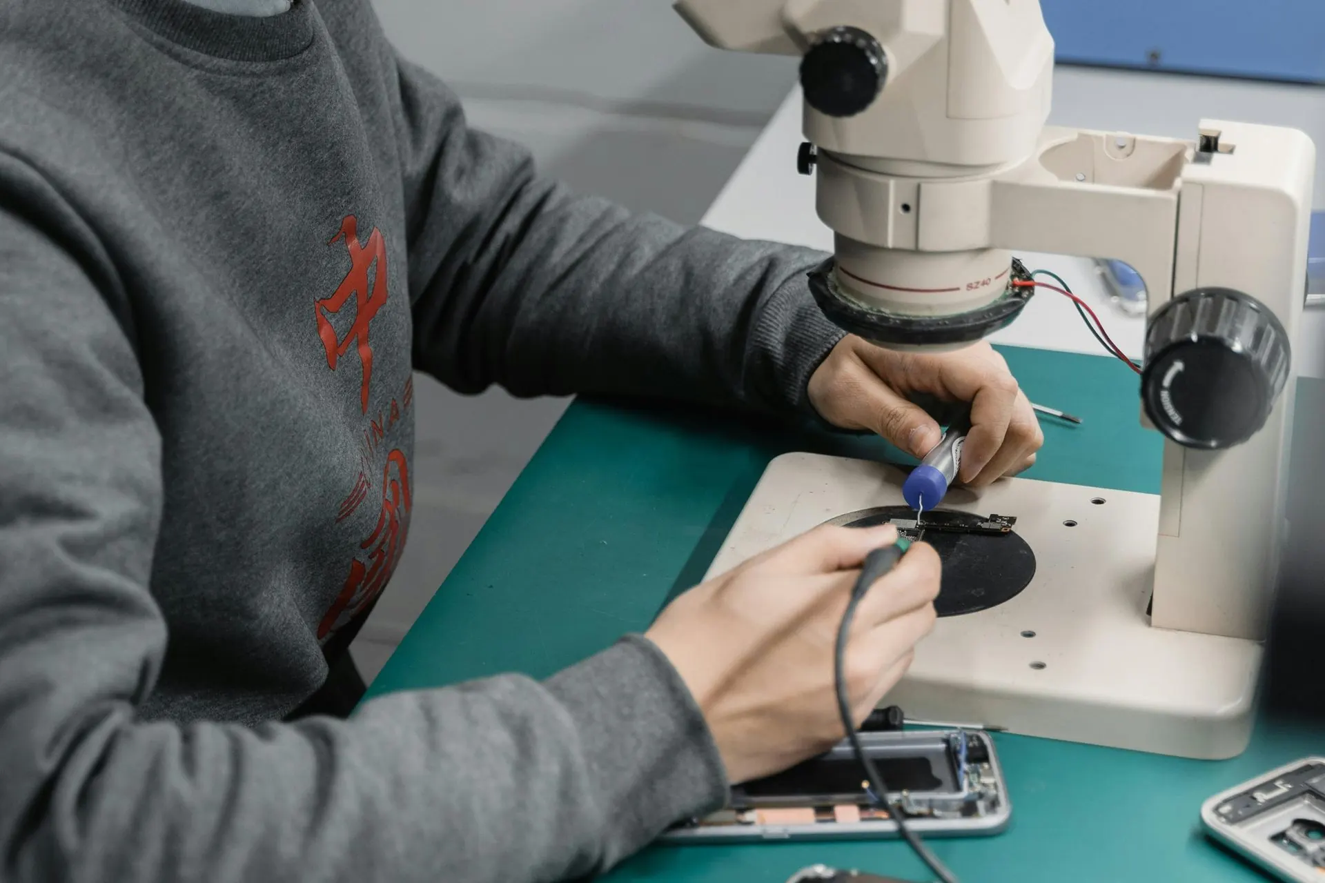Eine Person arbeitet unter einem Mikroskop an Elektronikgeräten; Hände benutzen Werkzeuge auf einer Telefonplatine.