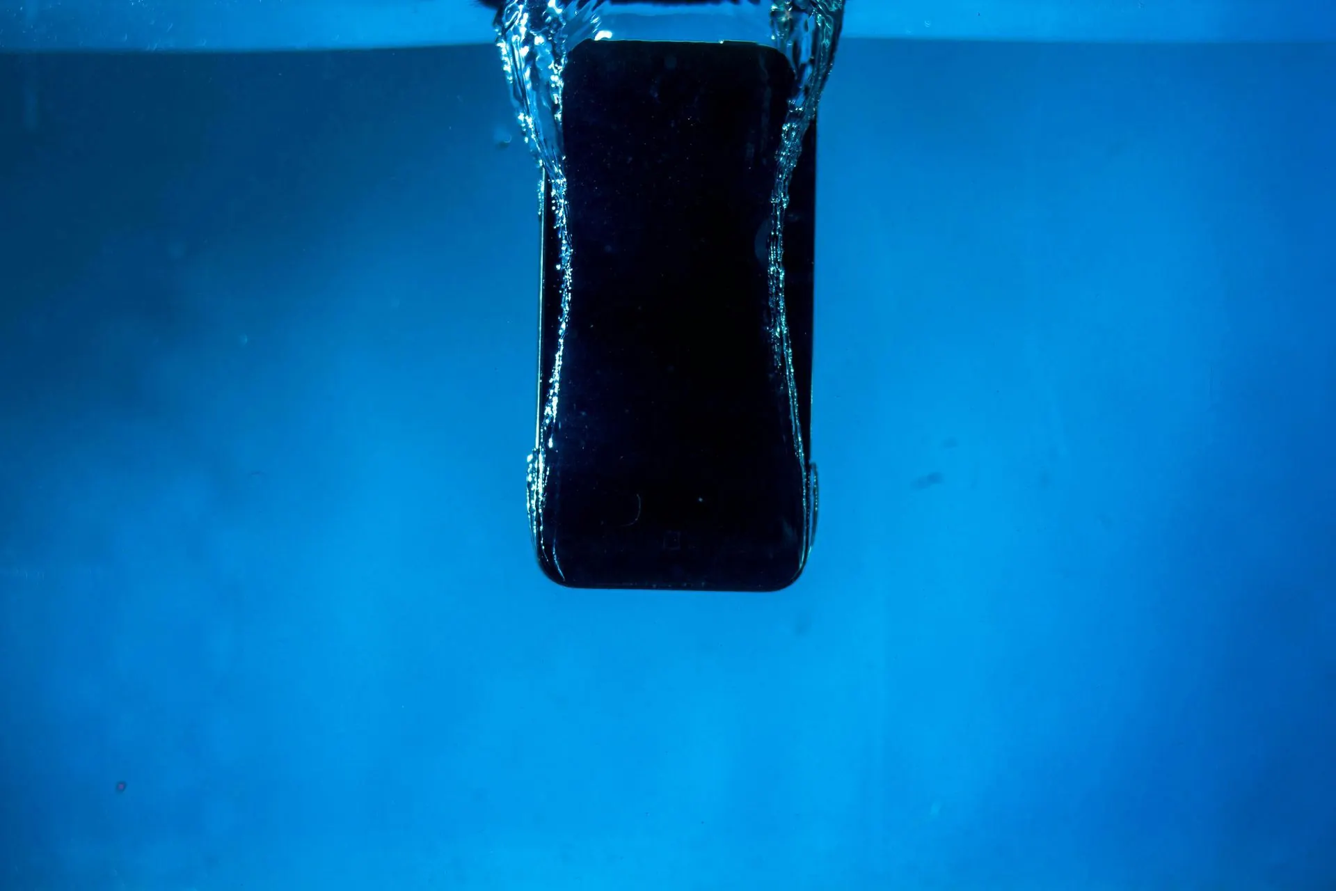 Ein schwarzes Handy wird in blaues Wasser getaucht und erzeugt Spritzer und Blasen.