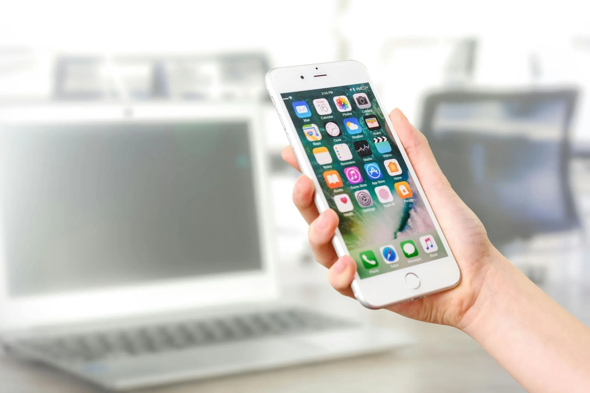 Eine Hand hält ein weißes Smartphone mit App-Symbolen; im Hintergrund ist ein verschwommener Laptop zu sehen.