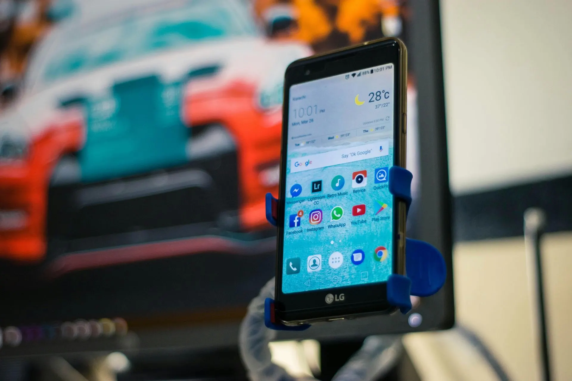 LG-Smartphone in einer blauen Halterung, auf dem Apps angezeigt werden, vor einem farbenfrohen Rennwagen-Hintergrund.